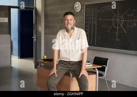 Der bärtige Lehrer mittleren Alters schaute im Klassenzimmer auf die Kamera. Glücklicher und engagierter Lehrer. Stockfoto