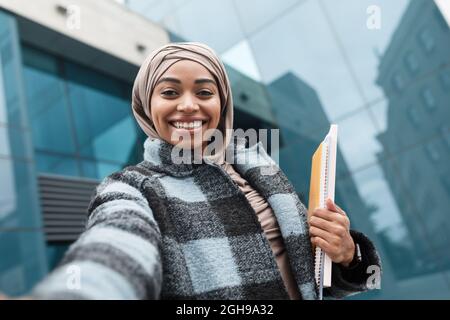 Zufriedene attraktive islamische afro amerikanische Studentin in Hijab und Mantel mit Büchern schaut auf Gadget Webcam Stockfoto