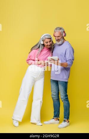In voller Länge ein glückliches Paar mittleren Alters und interracial in kabellosen Kopfhörern mit Gadgets auf Gelb Stockfoto