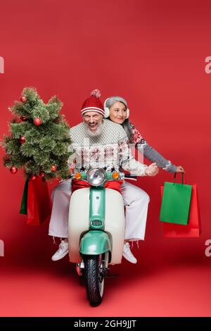 Glücklich mittleren Alters Mann hält weihnachtsbaum in der Nähe asiatische Frau, während Reiten Moped auf rot Stockfoto