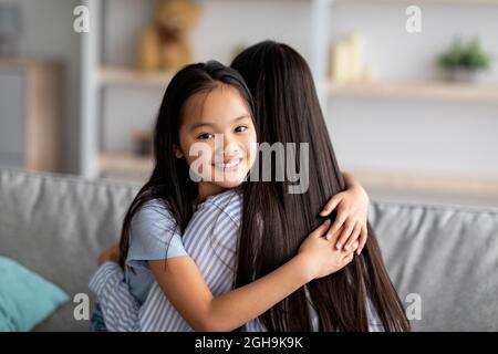 Glückliches asiatisches Schulmädchen umarmte ihre Mutter und lächelte vor der Kamera, saß zusammen auf dem Sofa, Nahaufnahme Porträt, Kopierraum Stockfoto