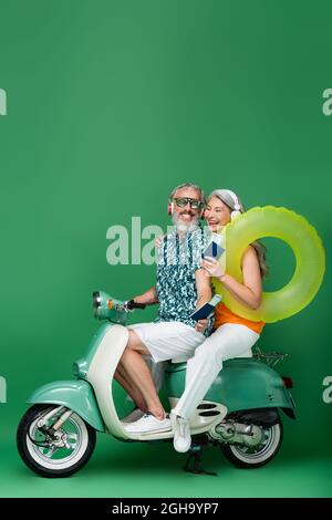 Ein glückliches und multiethnisches reifes Paar mit kabellosen Kopfhörern, die Pässe mit Flugtickets halten und auf Grün Motorroller fahren Stockfoto