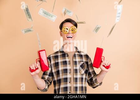 Foto von fröhlich verrückt Kerl halten Pistole werfen Schlag Geld tragen specs karierte Hemd isoliert beige Farbe Hintergrund Stockfoto