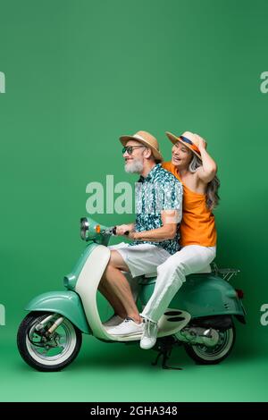 Positive und interracial mittleren Alters Paar in Sonnenhüten Reiten Motorroller auf grün Stockfoto