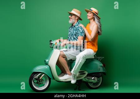 Glücklich interracial mittleren Alters Paar in Sonnenhüten Reiten Motorroller auf grün Stockfoto