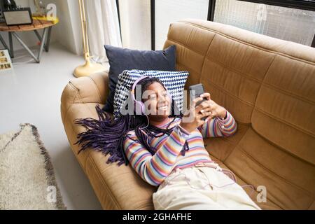 Lächelnde afroamerikanische Frau, die mit Kopfhörern Musik hört und mit dem Smartphone auf dem Sofa zu Hause entspannt.Konzept der Jugend und Freizeit. Stockfoto