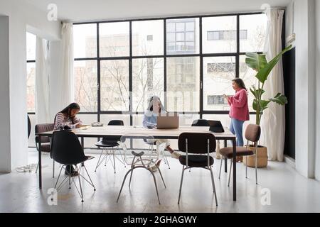 Im Büro. Drei Geschäftsfrauen arbeiten ohne Masken zusammen und halten dabei einen sicheren Abstand. Zwei von ihnen arbeiten am Schreibtisch und einer plaudert weiter Stockfoto