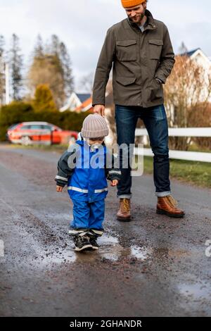 Ganzer Junge, der auf der Pfütze herumläuft, von Vater, der auf der Straße steht Stockfoto