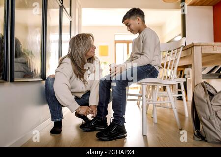 Mutter, die Schuhschnur eines autistischen Sohnes bindet, der auf einem Stuhl im Wohnzimmer sitzt Stockfoto