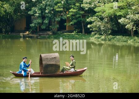 Lächelnd verheiratetes vietnamesisches Paar in traditionellen Kleidern und Kopfbedeckung Ruderboot in kleinem Teich Stockfoto