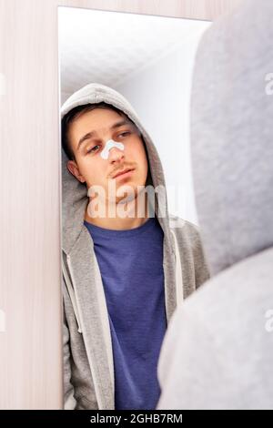 Trauriger junger Mann mit einem klebenden Gips auf der Nase in der Nähe des Spiegels im Haus Stockfoto