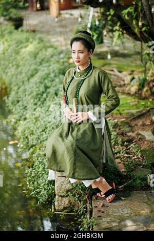 Ernst nachdenkliche junge Frau in traditioneller Tracht sitzt auf Stein am Teich und schaut auf das Wasser Stockfoto