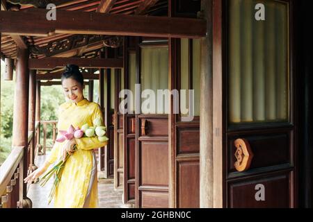 Schöne lächelnde junge Frau im Ao dai-Kleid, die auf der Veranda mit einem Haufen Lotusblumen steht Stockfoto