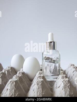 Auf einem Eierkarton steht eine Glasflasche mit Eiergesicht-Serum, in der Nähe befinden sich mehrere Eier auf dem weißen Hintergrund. Trendige natürliche Straffung. Stockfoto