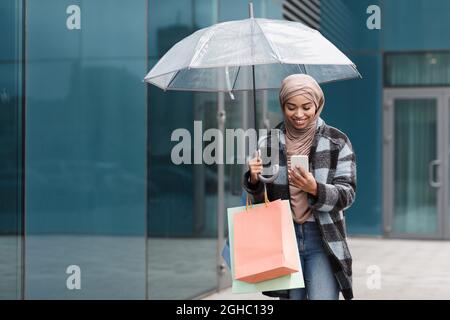 Fröhliche hübsche schwarze Frau in Hijab und Mantel mit Einkaufstaschen mit neuer Kleidung und Regenschirm Stockfoto