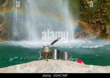 Kaffee im Freien zubereiten. Kaffeekocher auf dem Campinggasbrenner mit Wasserfall und Regenbogen im Hintergrund Stockfoto