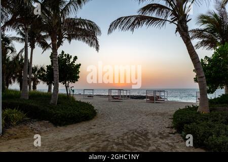 Leere Doppel-Sonnenliegen am Sandstrand in Dubai, mit orangefarbenem Sonnenuntergang im Hintergrund und ruhigem Meer, dunklen Silhouetten von Palmen. Stockfoto