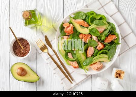 Lachs Spinat Avocado und grüner Pfeffer Salat auf einem weißen Teller mit goldener Gabel und Messer auf einem weißen Holztisch, mediterrane gesunde Ernährung, horizonta Stockfoto