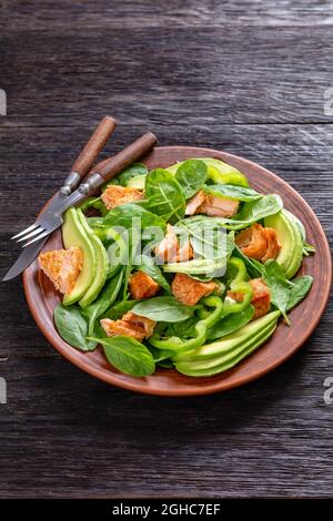 Lachs Spinat Avocado und grünen Pfeffer Salat auf einem braunen rustikalen Teller mit Gabel und Messer auf einem Mooreichen Tisch, mediterrane gesunde Ernährung, vertikale vie Stockfoto