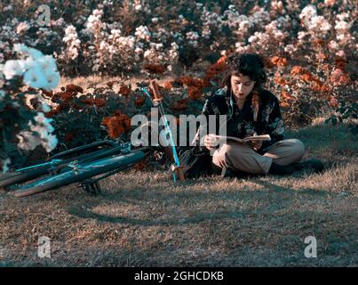 latina-Mädchen liest ein Buch auf dem Rasen des Parks mit Blumen im Hintergrund und einem Fahrrad neben ihr. Lesezeit horizontal Stockfoto