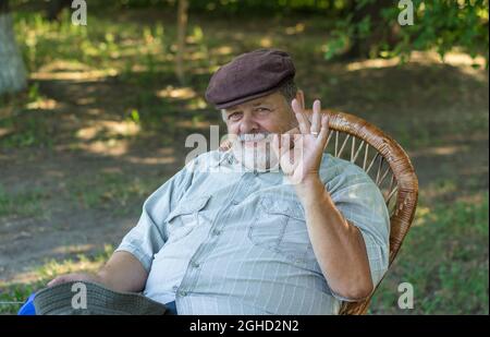 Außenportrait des kaukasischen älteren Mannes, der in einem Stuhl im Sommerpark sitzt und alles in Ordnung zeigt Stockfoto