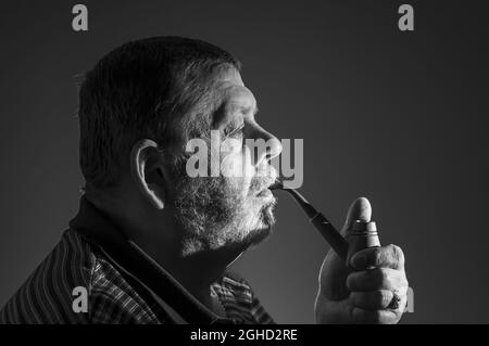 Schwarz-Weiß-Porträt eines kaukasischen bärtigen Mannes, der Tabakpfeife vor dunklem Hintergrund raucht Stockfoto