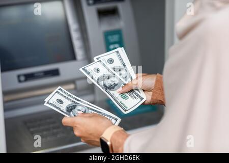 Hände einer nicht erkennbaren jungen arabischen Geschäftsfrau im Hijab zählen Dollar in der Nähe eines geldautomaten mit leerem Bildschirm Stockfoto