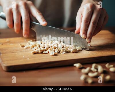 Weibliche Hände hacken Cashew. Nahaufnahme des Prozesses Zerkleinern roher, nicht gerösteter Cashew mit großem Messer auf Holzschneidebrett. Flacher Freiheitsgrad Selektiver Fokus Stockfoto