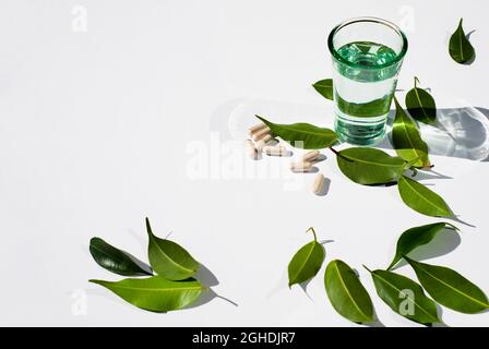 Glasbecher mit Wasser und Kapseln und Pillen auf weißem Hintergrund. Grüne Blätter. ... Alternative Medizin Kopieren Sie Leerzeichen für Text. Weißer Hintergrund. Stockfoto