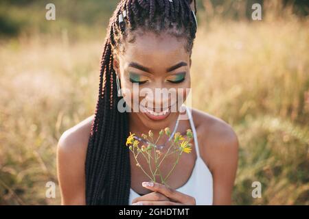 Schöne schwarze junge Frau im Park Porträt lange Zöpfe lächelnde Blume helle Make-up Nahaufnahme Stockfoto