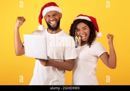 Weihnachtsangebot. Überglücklich schwarze Ehegatten in santa Hüte feiern Erfolg mit Laptop über gelbem Hintergrund Stockfoto