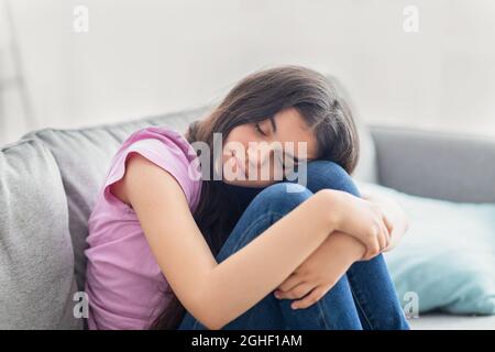 Unglückliches indisches Teenager-Mädchen, das mit geschlossenen Augen auf dem Sofa sitzt, ihre Knie umarmt, sich zu Hause depressiv oder verzweifelt fühlt Stockfoto