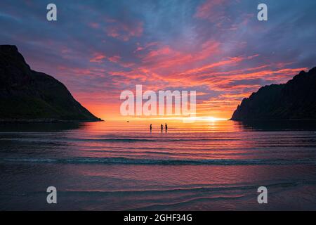 Drei Menschen, die während der Mitternachtssonne unter dem atemberaubenden bunten Himmel im Meer stehen, Ersfjord, Senja, Kreis Troms, Norwegen Stockfoto