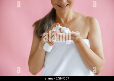 Lächelnde Dame mittleren Alters mit Handtuch drückt Körperpflegemittel auf die Hand auf rosa Hintergrund Stockfoto