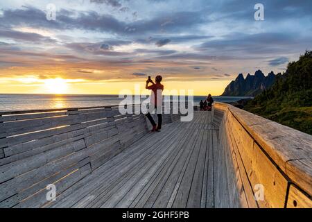 Person, die das Meer bei Sonnenuntergang mit dem Smartphone vom Pier am Aussichtspunkt Tungeneset, Senja, Kreis Troms, Norwegen, fotografiert Stockfoto