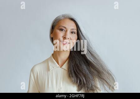 Lächelnde asiatische Frau mittleren Alters mit fließenden grauen Haaren steht auf hellem Hintergrund Stockfoto