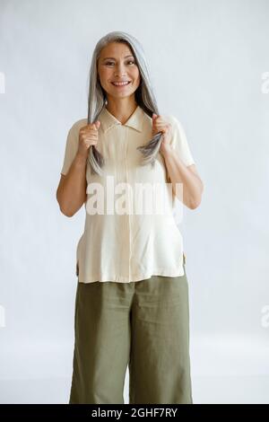 Lächelnde asiatische Frau mittleren Alters hält gesunde graue Haare auf hellem Hintergrund Stockfoto