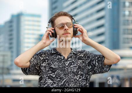 Ein junger Mann mit Kopfhörern und Sonnenbrillen hört vor dem Hintergrund moderner Gebäude Musik Stockfoto