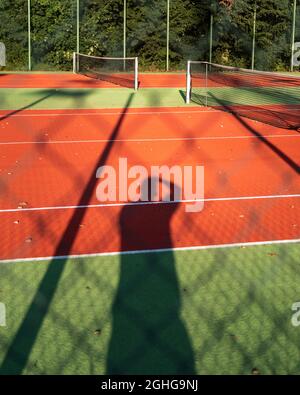 Schatten eines Mannes, der auf einem Tennisplatz Fotos macht. Rote Polyurethan-Oberfläche des Sportplatzes. Bäume im Hintergrund. Sonniger Tag. Stockfoto