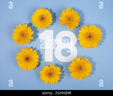 Nummer 18 umgeben von gelben Gerbera-Blüten auf blauem Hintergrund. Stockfoto