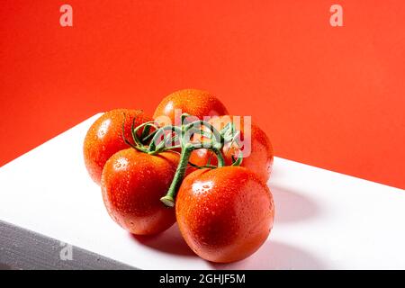 Foto eines Straußes von fünf nassen roten natürlichen Tomaten auf einem weißen Tisch und einem roten Hintergrund.das Foto wurde im horizontalen Format aufgenommen. Stockfoto