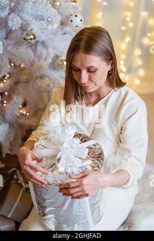 Porträt einer lächelnden kaukasischen Frau mit silberner Geschenkbox in der Nähe des Baumes. Eine junge Frau öffnet ihr Geschenk. Glückliches schönes Mädchen unter dem weihnachtsbaum. Stockfoto