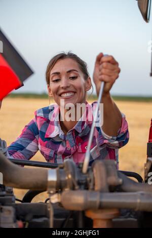 Hübsche junge Frau, die einen Schraubenzieher hält und versucht, den Traktor auf dem Feld zu reparieren Stockfoto