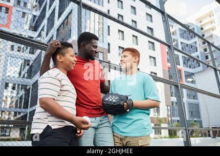 Lächelnder afroamerikanischer Vater plaudert nach einem Baseballspiel mit Söhnen auf dem Sportplatz gegen ein Wohngebäude Stockfoto