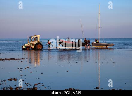 Segelboote/Schlauchboote werden bei Sonnenuntergang von einem Traktor auf Trolleys/Anhängern am Littlehampton Beach, West Sussex, Großbritannien, eingezogen Stockfoto