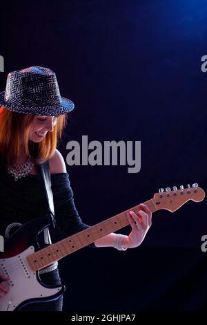 Hipster trendige junge Musikerin spielt Gitarre mit einem strahlenden Lächeln der Freude Seite beleuchtet über einem dunkelblauen Hintergrund mit Copyspace Stockfoto