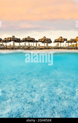 (Selektiver Fokus) Split Shot, über dem Bild. Kristallklares, türkisfarbenes Wasser und einige Sonnenschirme, die während eines atemberaubenden Sonnenaufgangs beleuchtet werden. Stockfoto