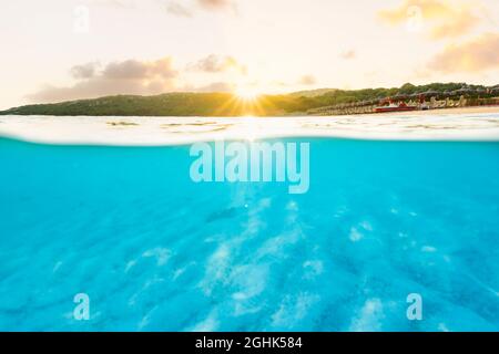 (Selektiver Fokus) Split Shot, über dem Bild. Kristallklares, türkisfarbenes Wasser und einige Sonnenschirme, die während eines atemberaubenden Sonnenaufgangs beleuchtet werden. Stockfoto