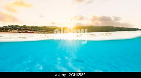 (Selektiver Fokus) Split Shot, über dem Bild. Kristallklares, türkisfarbenes Wasser und einige Sonnenschirme, die während eines atemberaubenden Sonnenaufgangs beleuchtet werden. Stockfoto