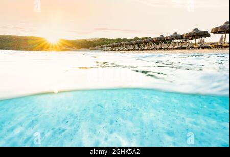 (Selektiver Fokus) Split Shot, über dem Bild. Kristallklares, türkisfarbenes Wasser und einige Sonnenschirme, die während eines atemberaubenden Sonnenaufgangs beleuchtet werden. Stockfoto